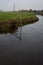 Stream of water in the countryside and a mooring pole by the shore with its reflection casted in the water on a cloudy day