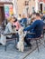 Street cafe, family with dogs resting. Man patting dog`s head, dog standing on hind legs, other dog lies on woman hands