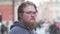 Street portrait of a serious, fair-haired 25-30-year-old man with glasses and a beard against the background of city buildings and