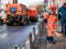 Street worker sweeping the sidewalk in an orange uniform. In the background, special watering machines are visible for cleaning s