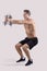 Strength workout. Powerful young sportsman with dumbbells doing squats on light studio background, full length portrait