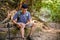 A stressed Asian male trekker sits on a wooden log and holds his knee, suffering from knee pain