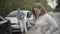 Stressed desperate young woman looking at crashed car with blurred man examining damage at the background. Hopeless