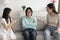 Stressed mature Asian woman sitting on couch while adult daughter and granddaughter arguing with her at home