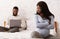 Stressed pregnant woman sitting on bed while husband using laptop