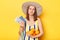 Stressed woman on resort dressed in striped swimwear and straw hat posing isolated over yellow background holding fruit and
