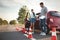 Stressed young woman in car near instructor and fallen traffic cones outdoors. Failed driving school exam