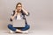 Stressed young woman sitting on the floor with crossed legs and using laptop on gray background. Using phone