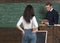 Strict professor in glasses reading a book at rostrum. Female brunette student holding hands in rear jeans pockets while