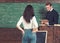 Strict professor in glasses reading a book at rostrum. Female brunette student holding hands in rear jeans pockets while