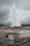 Strokkur geyser erupting in Iceland with spectators standing nearby in a cloudy day