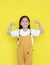 Strong asian little kid girl raising hands up and smiling. Portrait of happy child in dungarees isolated on yellow background