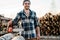 Strong bearded lumberjack wearing plaid shirt hold chainsaw in hand on background of sawmill