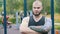 A strong bearded man with tattoos standing on the sports ground outdoors - looking in the camera with his arms crossed