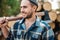 Strong bearded woodcutter holds axe on his shoulder on warehouse of logs