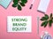 STRONG BRAND EQUITY is written in green on a white notepad on a pink background surrounded by notepads, pens, white alarm clock