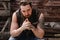 Strong brutal man with a beard and tattoos on his hands dressed in leather vest and jeans sits on a wooden wall