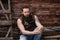 Strong brutal man with a beard and tattoos on his hands dressed in leather vest and jeans sits on a wooden wall