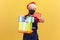 Strong courier in medical protective mask, uniform and santa claus hat holding many gift boxes and showing sale coupon, xmas sales