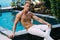 Strong handsome shirtless man in white pants sits on pool in garden. Fitness model posing on camera