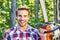 Strong lumberjack with chainsaw in a plaid shirt. Lumberjack worker walking in the forest with chainsaw. Woodcutter with