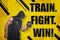 Strong muscular young man in black sportswear with a dumbbell and `TRAIN. FIGHT. WIN ` sign on yellow background