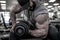 Strong young bearded man in cap lifting heavy weight dumbbell on muscle biceps sitting with great effort and pain during hard