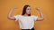 Strong young woman showing muscles, looking happy isolated on orange wall