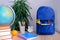 Student home office table with blue schoolbag, books, orange, colored notebooks, pencils in glass, chalk board, globe, white alarm