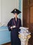 Student mannequin, inside a library, dressed as a graduate with a black academic cap and gown, holding a diploma in hand.