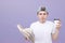 Student in a white T-shirt stands with a book and a cup of coffee ion a purple pacing background and looks at the camera