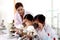 Students and teacher in lab coat have fun together, two little children students look through microscope and learn science