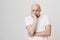 Studio portrait of gloomy bald european male model showing boredom while leaning head on hand and standing over gray