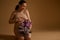 Studio portrait of gorgeous pregnant woman putting her hand on belly, holding bunch of purple lilacs, isolated on beige