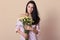 Studio portrait of miling Caucasian woman wearing pink dress, holding bouquet of flowers  over beige background,