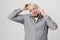 Studio portrait of pleased and happy bald adult male scratching his head while talking on smartphone, standing over gray