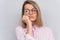 Studio portrait of sad blonde female student wearing pink shirt and round spectacles, sitting with unhappy boring expression on