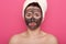 Studio shopt of unhappy beautiful young woman with black clay face mask makes acne therapy, wearing white towel on head, posing