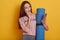 Studio shot of astonished dark haired girl with perfect body, holding in hands yoga mat and modern smart phone, reading shocked