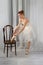 Studio shot of a calm beautiful ballerina in a white airy classic dress put her foot on a vienna chair and tying ribbons on pink