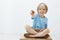 Studio shot of carefree happy blond child being entertained, sitting with crossed feet and pointing aside while smiling