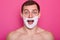 Studio shot of cheerful surprised man with shaving foam on his face looks directly at camera with open mouth, has astonished