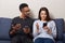 Studio shot of couple sitting together on comfortable sofa in apartment and using mobile phone, looking on screen, dark skinned