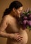 Studio shot of a delightful happy adult pregnant woman with bouquet of lilacs, touching her belly over beige background