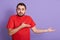 Studio shot of discontented man wearing redcasual t shirt, spreading his hands aside, posing against purple background, bearded