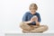 Studio shot of frustrated intense cute young boy with blond hair in casual outfit, sitting on crossed feet, bending