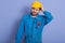 Studio shot of handsome young bearded Caucasian mechanic wearing blue work clothes and yellow hard hat, stands touching his head,