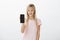 Studio shot of intrigued neat blond girl in pink t-shirt, smiling curiously and showing black smartphone at camera