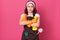 Studio shot of serious housewife wears orange rubber gloves, shirt and brown apron, holds detergent bottles and duster, being