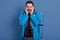 Studio shot of unshaven guy wearing blue jacket, holding mat and binoculars, touching his cheeks with both hands, traveller does